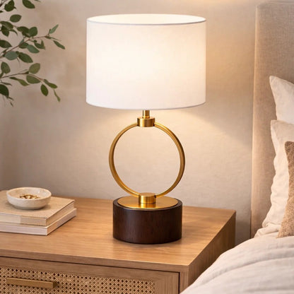 Ring table lamp with circular metal frame, walnut wood base and white linen drum shade on a bedside nightstand